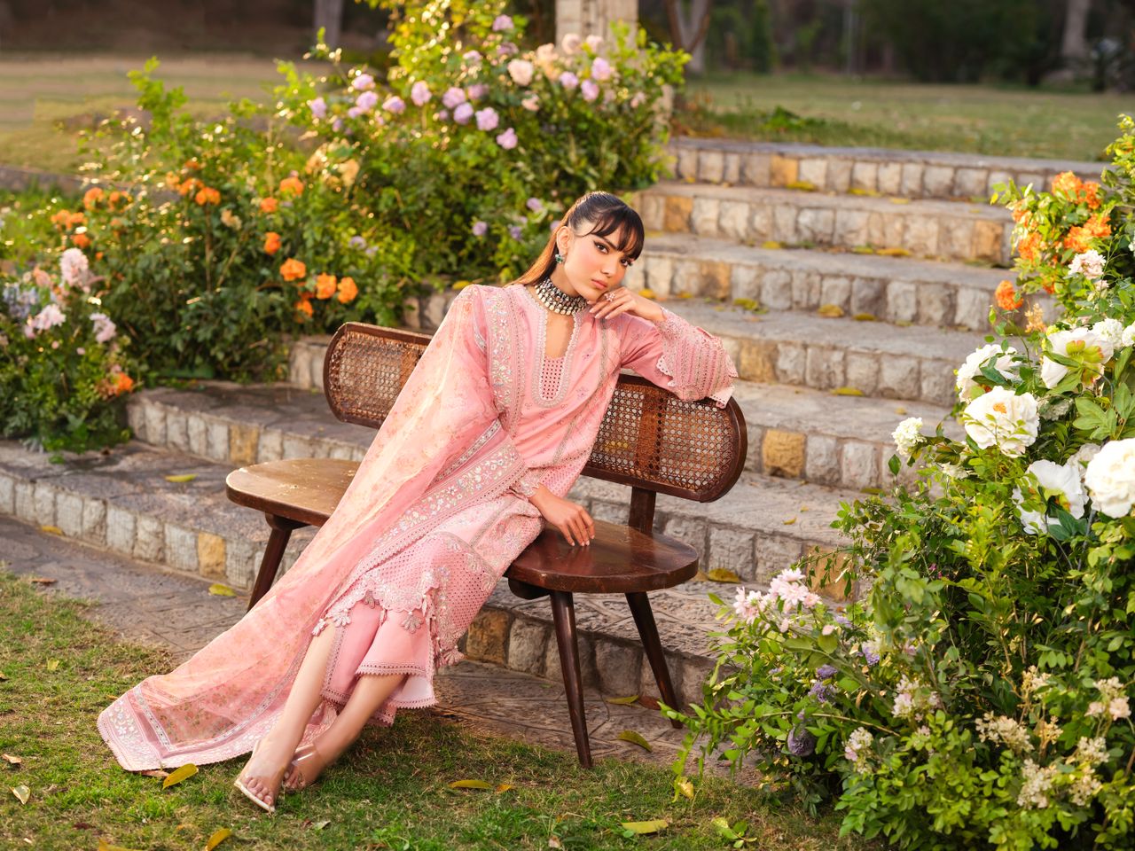 services-03 A fashionable model in a pink traditional dress seated outdoors among vibrant flowers.